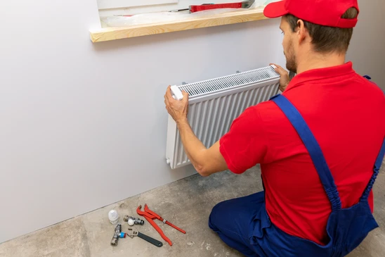 Intérieur résidentiel avec technicien en combinaison rouge et bleue installant un radiateur blanc à ailettes sur mur gris