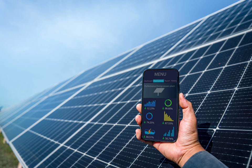 Scène extérieure avec main tenant smartphone affichant interface de monitoring énergétique, arrière-plan de panneaux solaires photovoltaïques sous ciel bleu