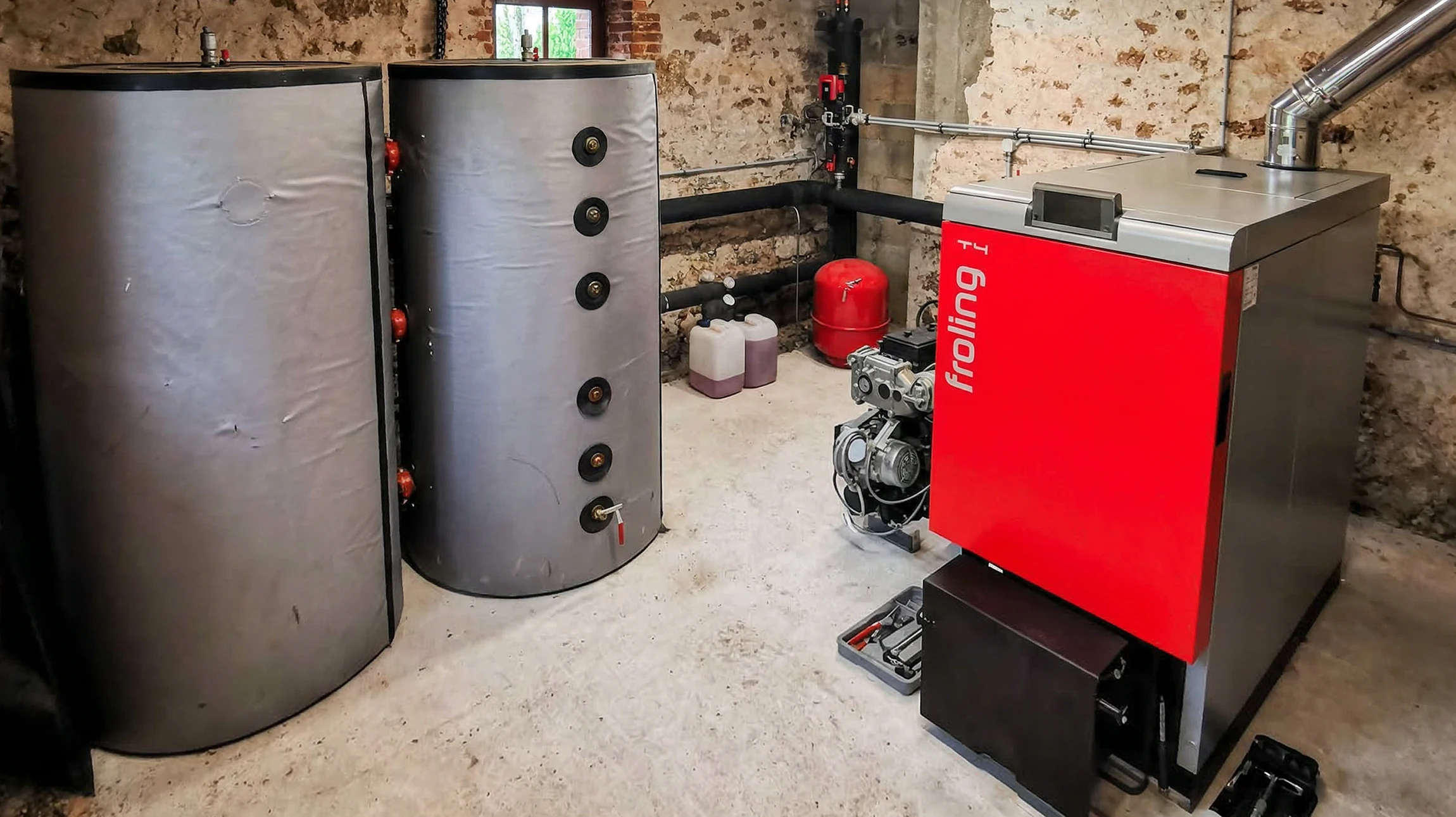 Photographie professionnelle d'une installation de chaudière gaz ou fioul moderne dans une chaufferie résidentielle. Le technicien qualifié porte des vêtements professionnels et effectue les raccordements finaux ou l'inspection de l'unité. L'image montre la chaudière fixée au mur ou sur un support, avec ses tuyauteries de raccordement, ses connexions et ses éléments de contrôle visibles. L'environnement est une chaufferie ou sous-sol résidentiel bien éclairé. La mise au point est nette sur le travail et la qualité de l'installation, mettant en évidence l'expertise professionnelle.