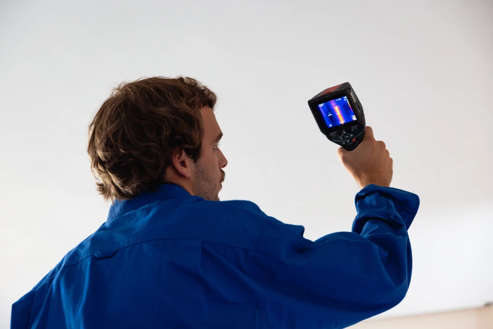 Plombier professionnel en intérieur, équipé d'un détecteur acoustique ou thermique, inspectant un mur ou une canalisation encastrée dans une habitation. Le technicien porte une tenue professionnelle de plombier (bleu de travail ou uniforme avec logo), tenant l'appareil de détection dirigé vers une zone de conduite d'eau. L'arrière-plan montre l'intérieur d'une maison contemporaine, avec des murs neutres et un environnement domestique clair. L'image doit suggérer la précision, la technologie et la réactivité d'intervention à domicile.