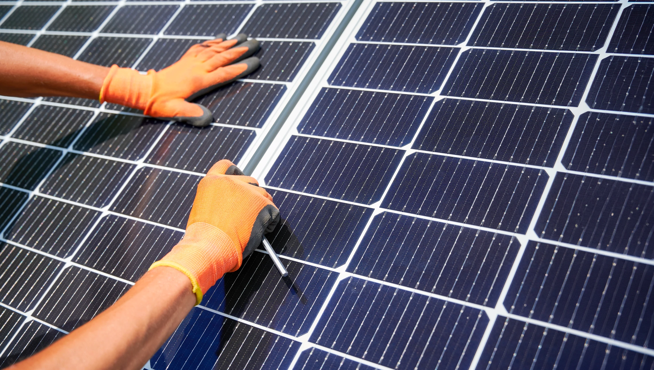 Ouvrier en tenue de sécurité haute visibilité et casque jaune installant des panneaux solaires photovoltaïques sur toiture résidentielle en bardeaux rouges, ciel bleu dégagé